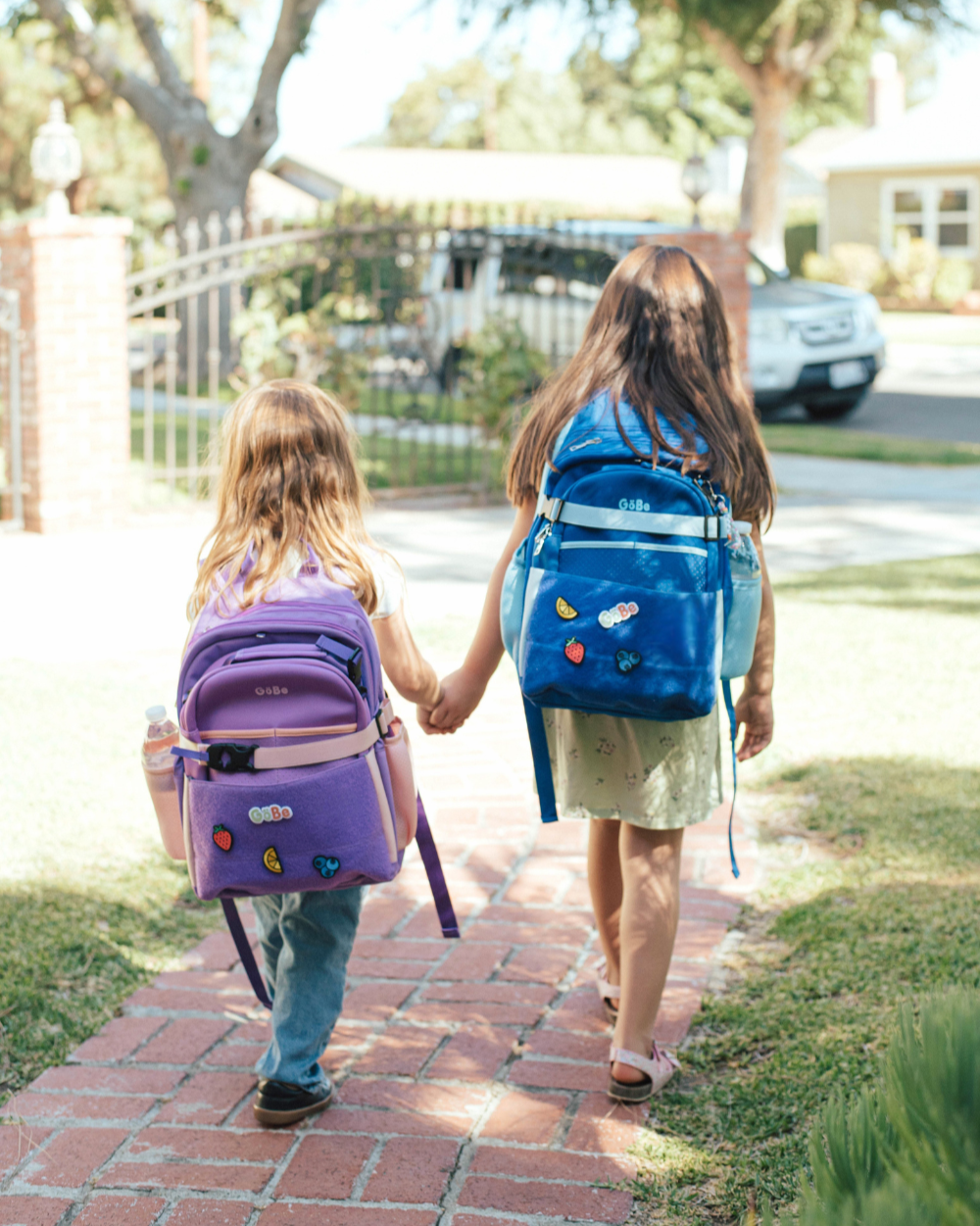 Two children walking down a sidewalk holding hands, each with a backpack.