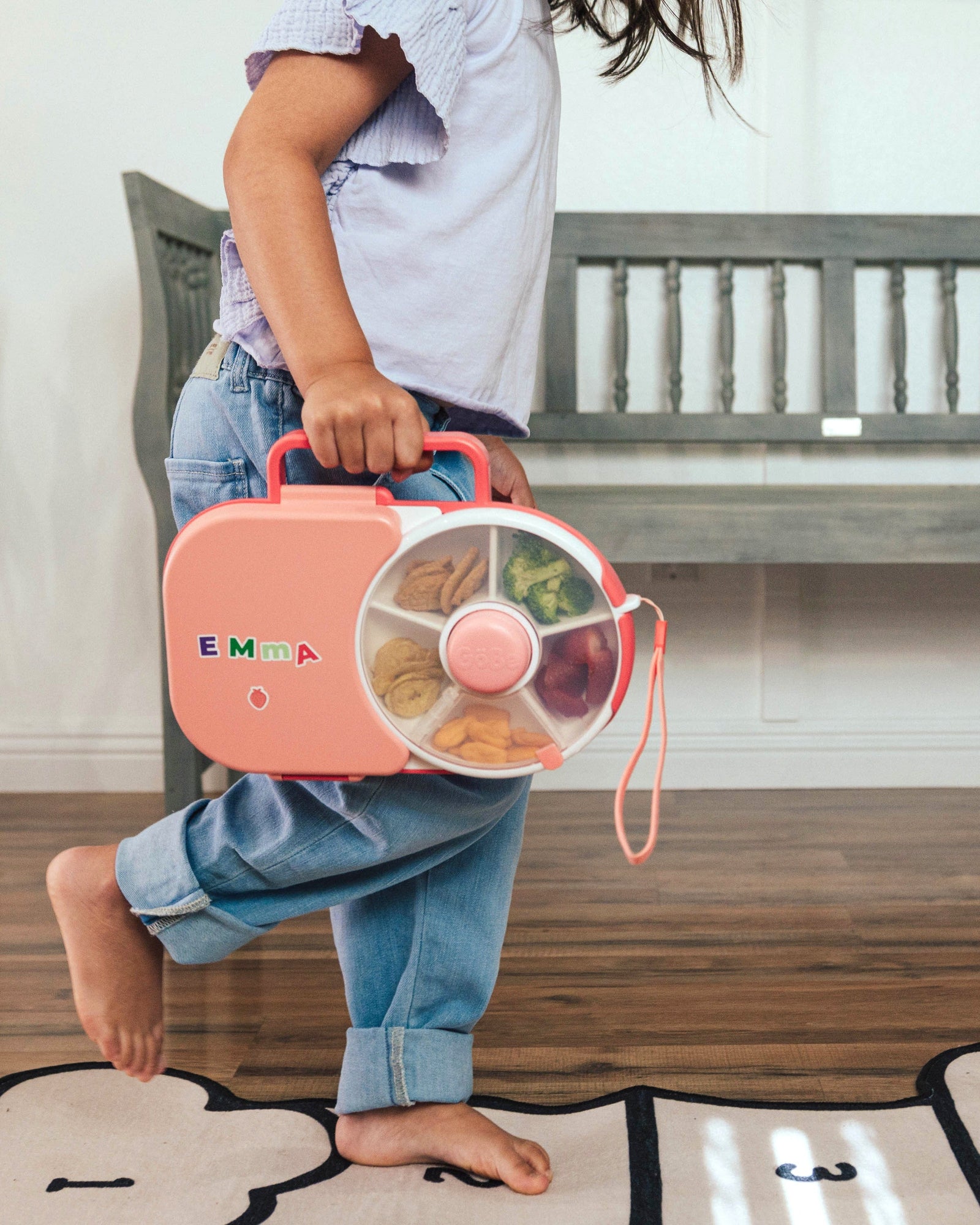 Child holding a pink GoBe Lunchbox with food items on a wooden floor.